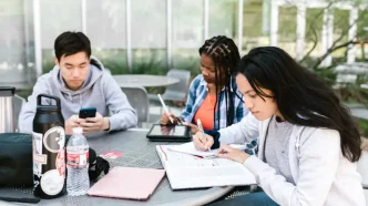 Tres estudiantes universitarios sentados en una mesa exterior trabajando con libros y dispositivos digitales.