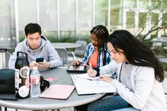 Tres estudiantes universitarios sentados en una mesa exterior trabajando con libros y dispositivos digitales.