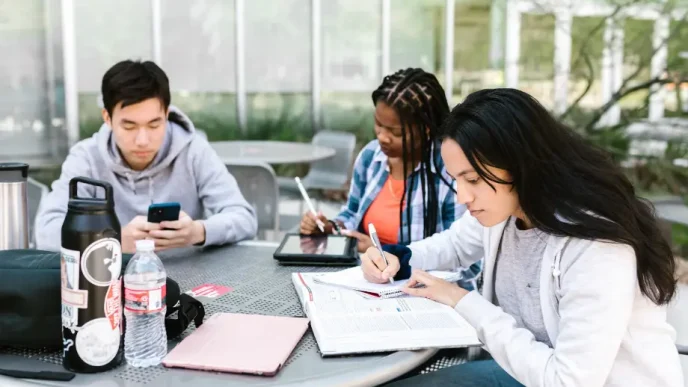 Tres estudiantes universitarios sentados en una mesa exterior trabajando con libros y dispositivos digitales.