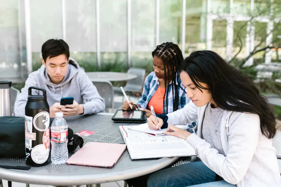Tres estudiantes universitarios sentados en una mesa exterior trabajando con libros y dispositivos digitales.