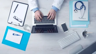 Médico escribiendo en un ordenador portátil en su consulta junto a un estetoscopio y documentos médicos.