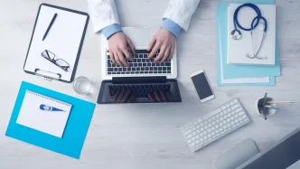 Médico escribiendo en un ordenador portátil en su consulta junto a un estetoscopio y documentos médicos.