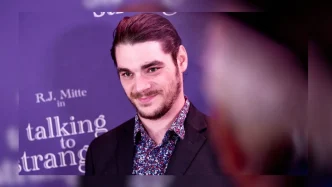 El actor RJ Mitte sonriendo en una alfombra roja con traje oscuro y camisa estampada.