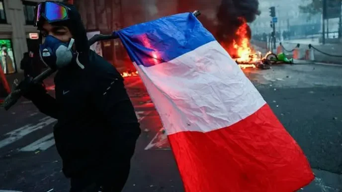 Manifestante con máscara de gas y la bandera de Francia durante unos disturbios urbanos con fuego de fondo.