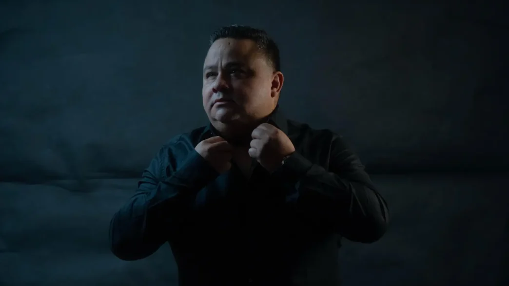 Retrato del mentor ecuatoriano John Arguello ajustándose el cuello de una camisa negra sobre un fondo oscuro.