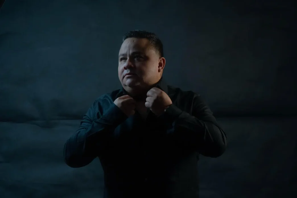 Retrato del mentor ecuatoriano John Arguello ajustándose el cuello de una camisa negra sobre un fondo oscuro.