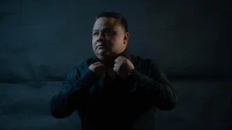 Retrato del mentor ecuatoriano John Arguello ajustándose el cuello de una camisa negra sobre un fondo oscuro.
