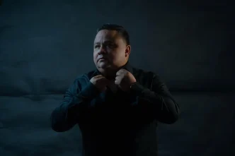 Retrato del mentor ecuatoriano John Arguello ajustándose el cuello de una camisa negra sobre un fondo oscuro.