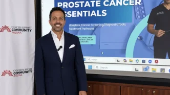 El médico oncólogo británico Ajay Bhatnagar posa en traje frente a una pantalla sobre cáncer de próstata.