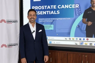 El médico oncólogo británico Ajay Bhatnagar posa en traje frente a una pantalla sobre cáncer de próstata.